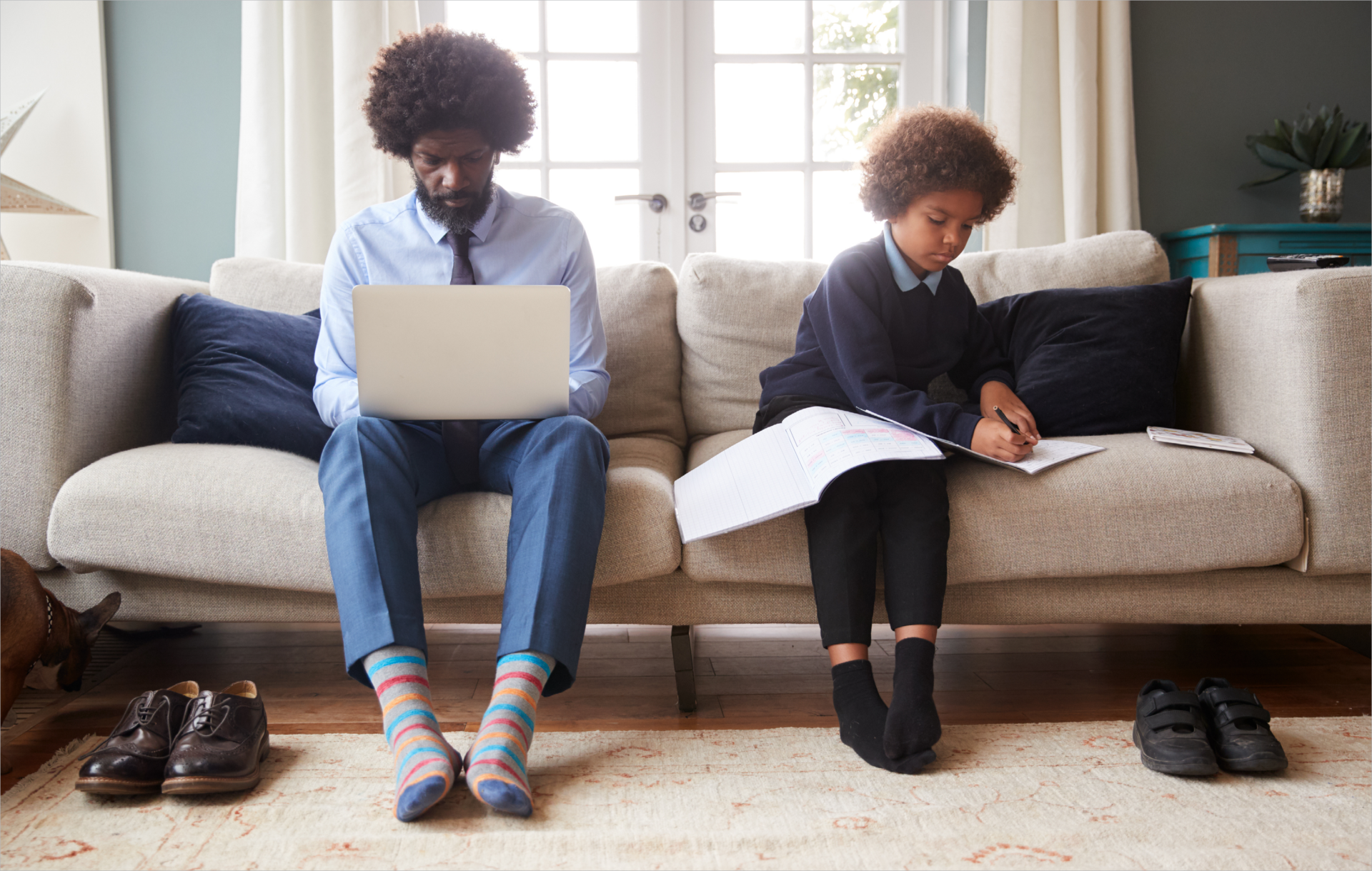 Two children sit at a table with Chromebooks, facing each other and talking, in a classroom with books and storage bins in the background.