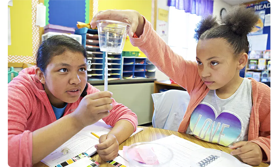 Two students engage in a science experiment in a classroom using high quality instructional materials, one holding a beaker above a petri dish as the other observes intently.