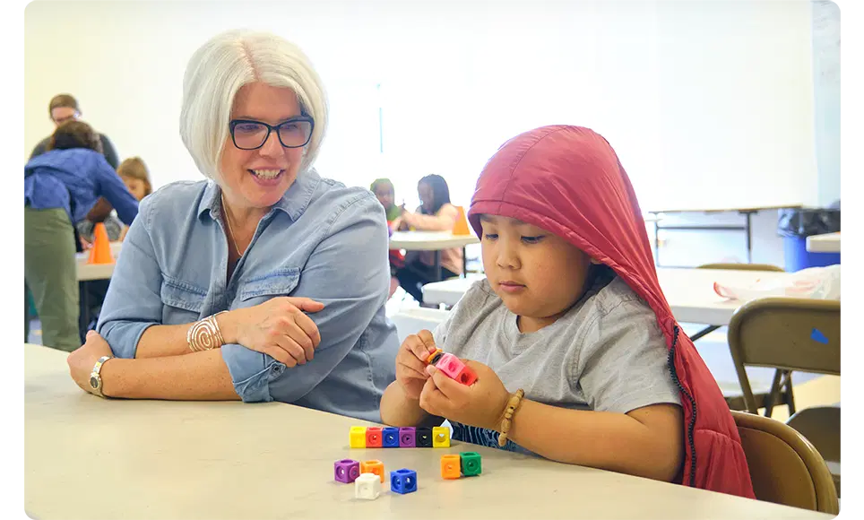 A woman in a blue shirt and glasses smiles while observing a young girl in a red headscarf playing with colorful blocks at a classroom table with High Quality Instructional Materials.