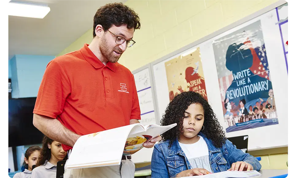 A teacher in a red shirt and glasses reading a book to a young girl in a classroom, with High Quality Instructional Materials on the wall behind them.