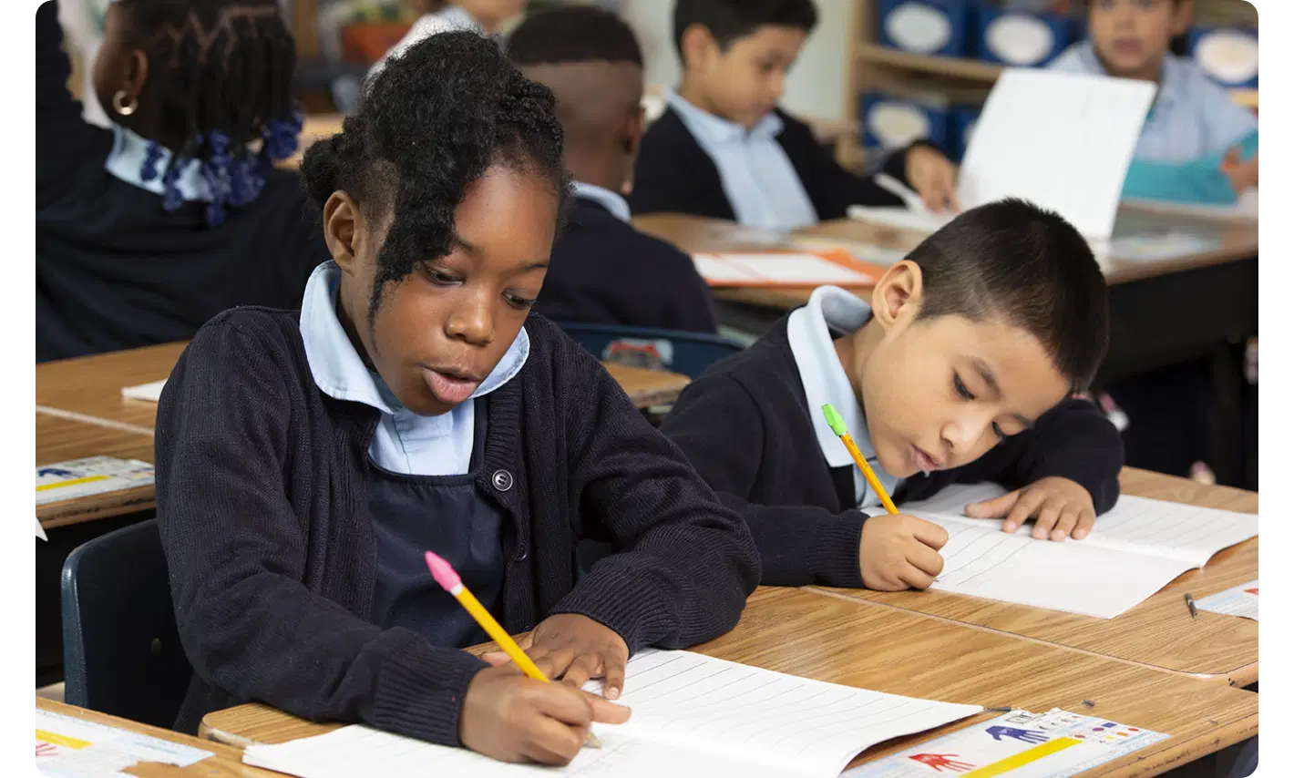 Two students in school uniforms writing in notebooks at their desks in an Amplify education classroom, with other classmates in the background.