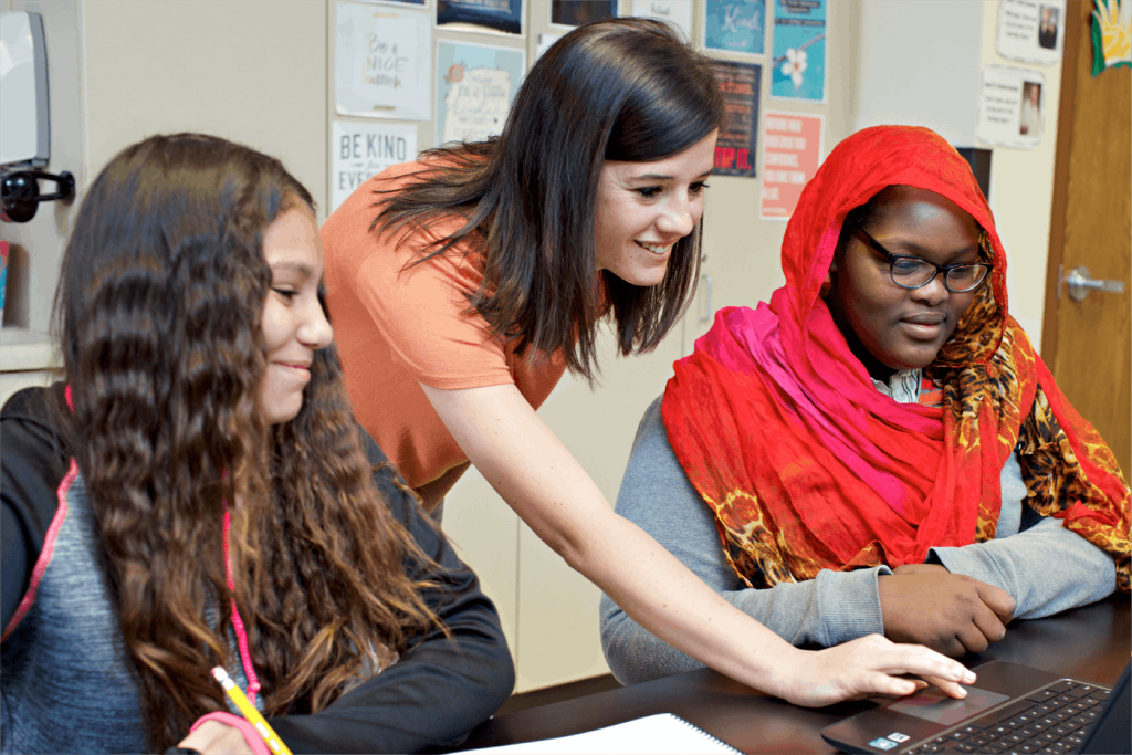 A teacher assists two students with a laptop in a classroom, one wearing a hijab, conveying a supportive educational environment.