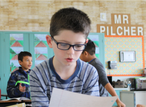 A boy wearing glasses reads a sheet of paper in a classroom. Other students are visible in the background. A sign reads "Mr Pilcher.