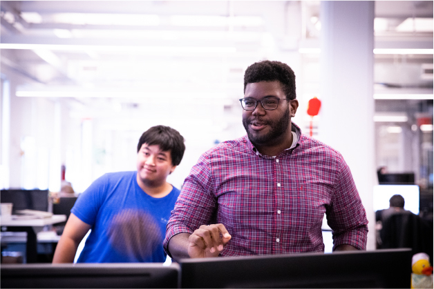Two men, one wearing glasses, using computers in a busy office setting with blurred background.