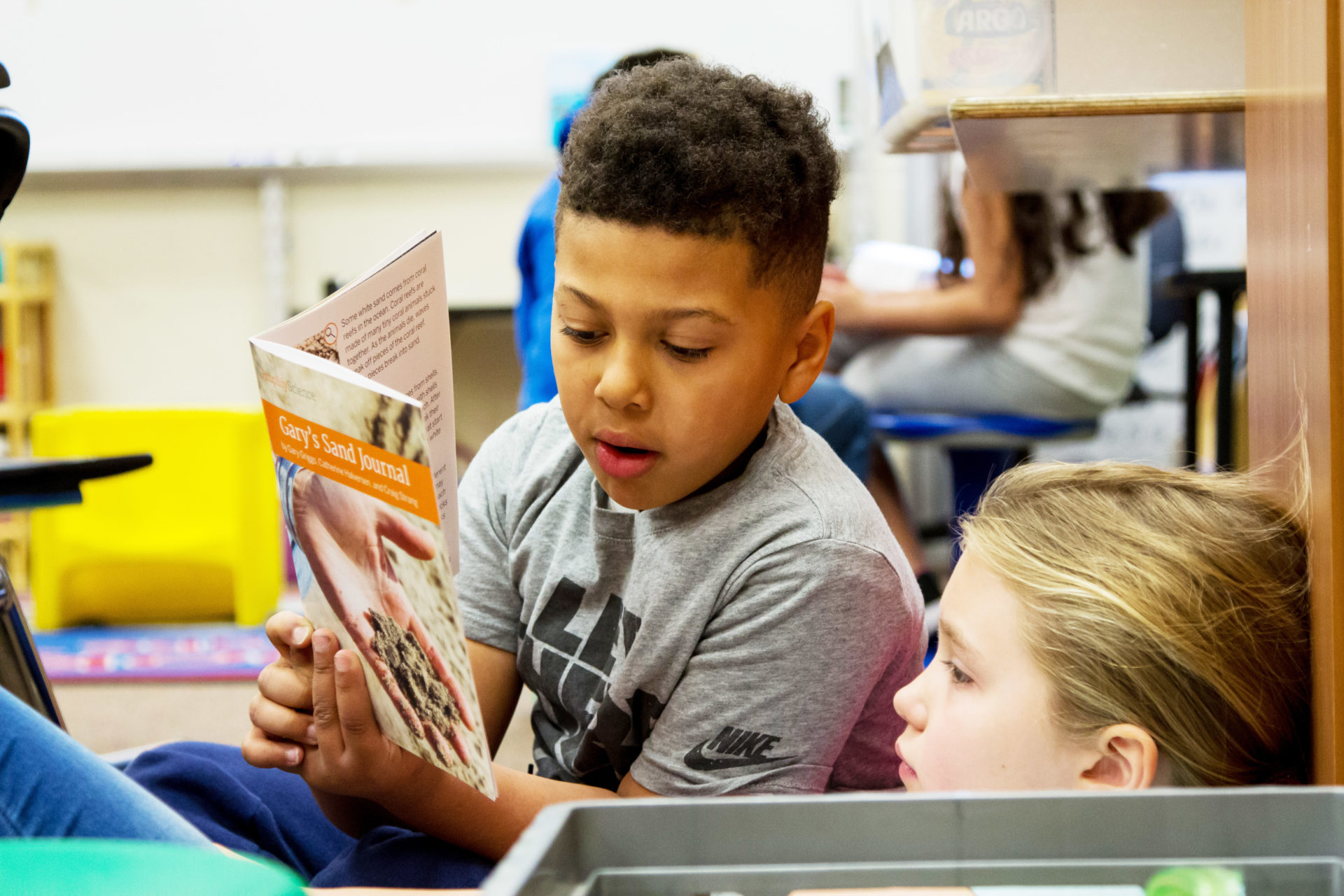 A young boy and girl sitting in a classroom, reading a book together with focused expressions.