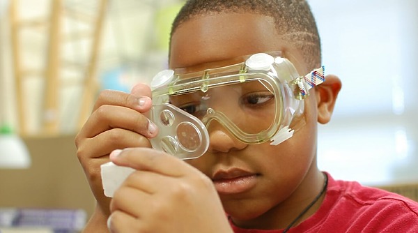 A child wearing safety goggles closely examines an object through a magnifying glass in a classroom setting.