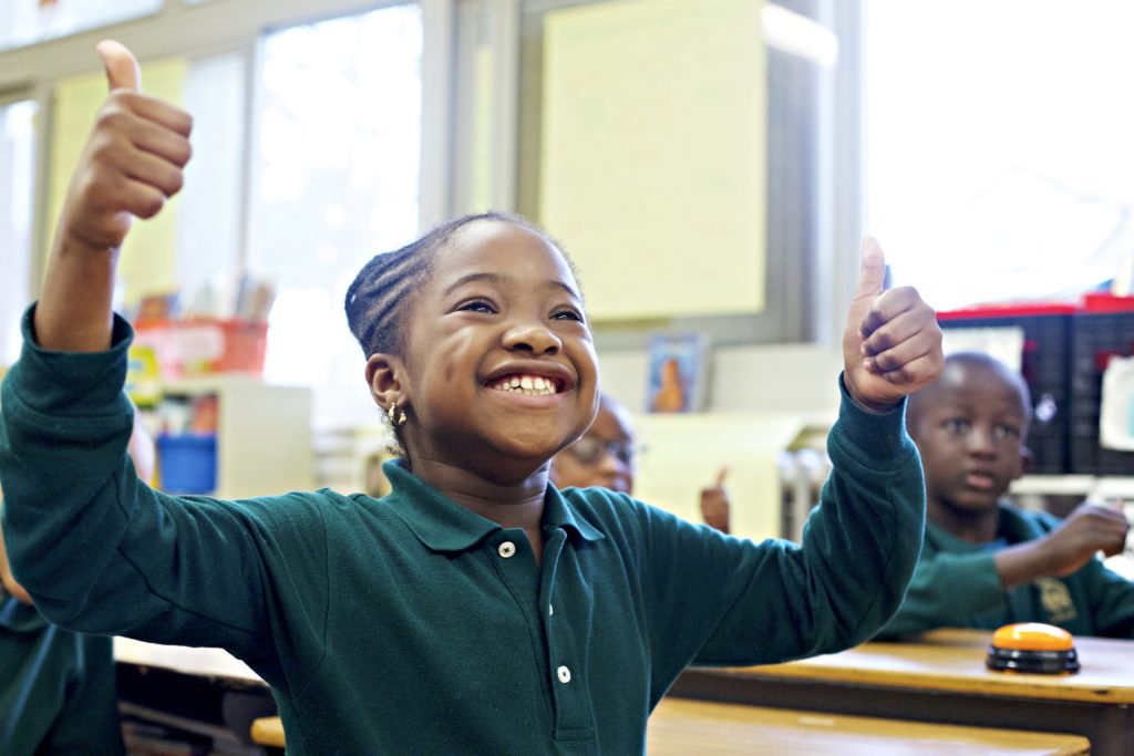 A young girl in a classroom gives a thumbs up with both hands, smiling broadly, while other students look on in the background.