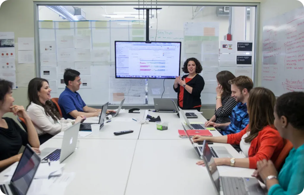 A group of people sit around a conference table with laptops. A woman stands at the front, presenting information displayed on a large screen.