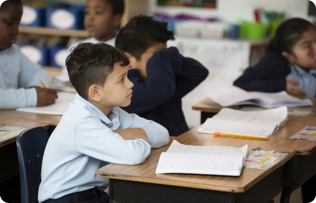 Children sitting at their desks in a classroom, listening attentively. Open notebooks and pencils are on the desks.