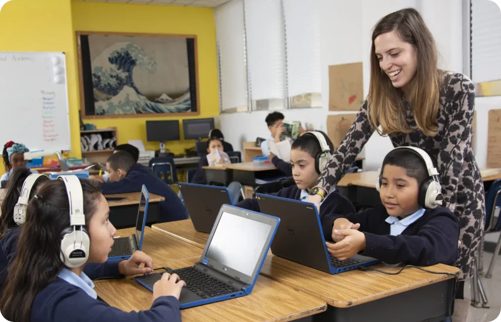 Students in a classroom work on laptops with headphones on, while a teacher stands by their side, smiling and helping them.