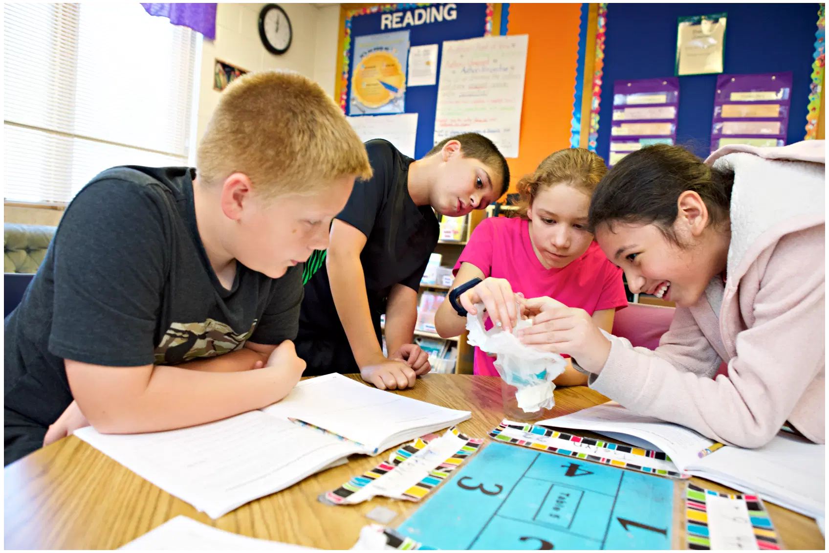 Four middle school science students gather around a desk, engaged in an interactive group project, investigating an object together in a colorful classroom setting.