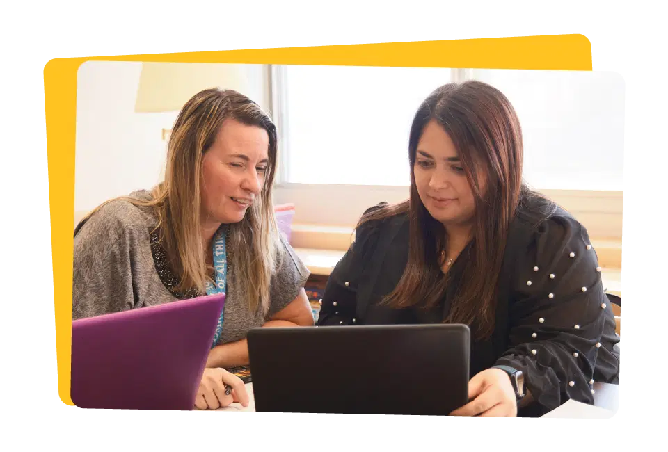 Two women working together on laptops at a table in an office environment.