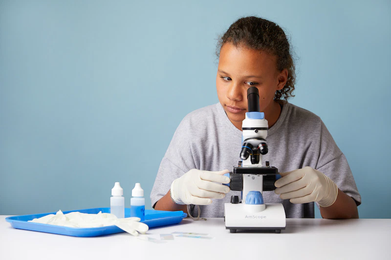 A young person wearing gloves looks through a microscope at a table with laboratory supplies, including bottles, slides, and a tray, against a plain blue background.
