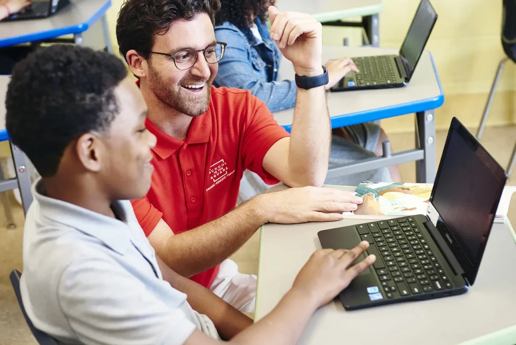 A teacher in a red shirt and glasses is smiling and assisting a student at a desk with a laptop in a classroom. Other students with laptops are in the background.