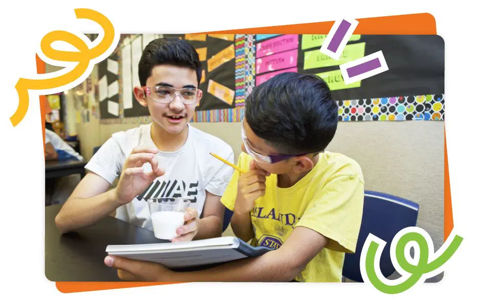 Two boys sit at a classroom desk; one holds a notebook and pencil while the other gestures and holds a plastic cup. Both wear safety glasses.