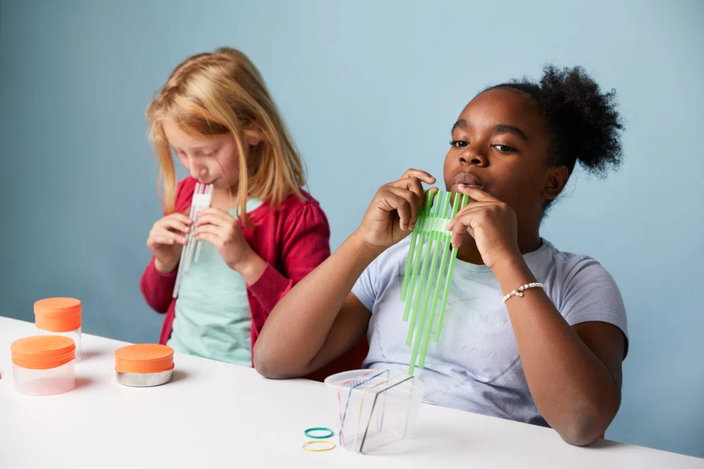 Two children sit at a table blowing into plastic straws held together, likely making musical sounds, with containers and rubber bands on the table.