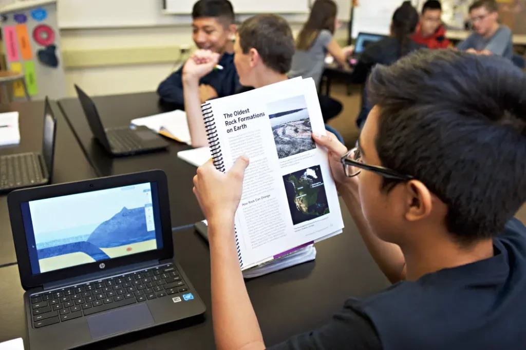 A student reviews a science textbook and looks at a laptop in a classroom, while other students work together on their science projects in the background.