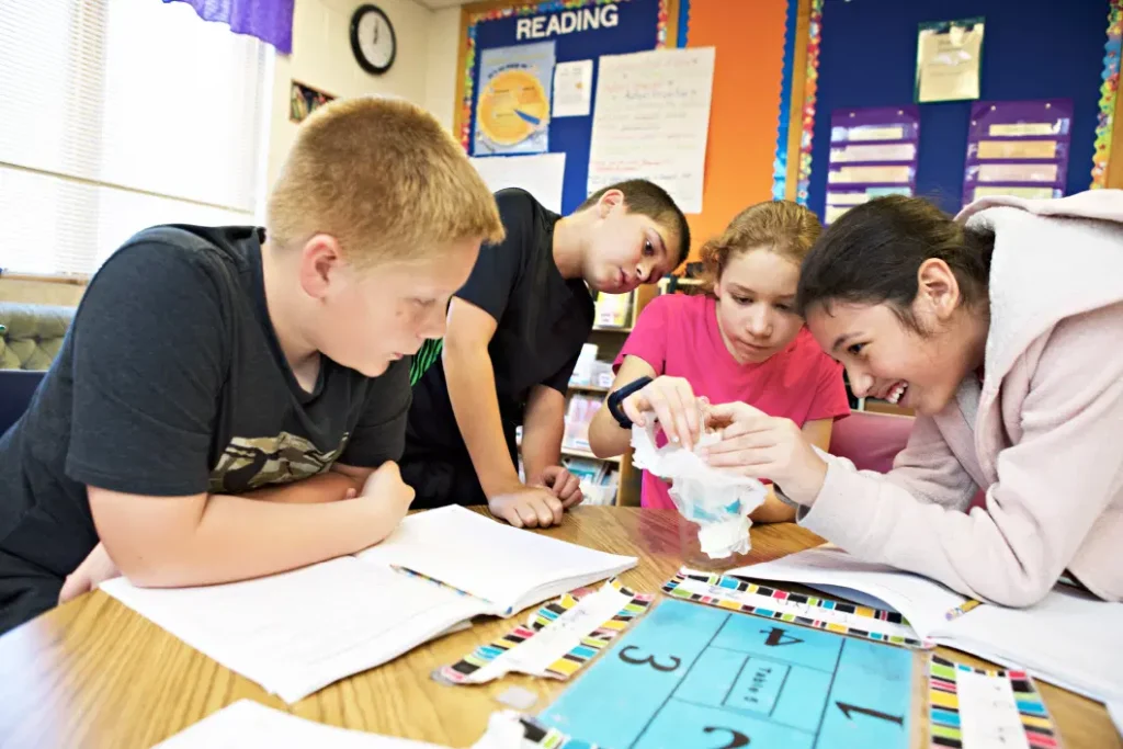 Four students work together at a classroom table, examining a science project while surrounded by notebooks and educational materials.