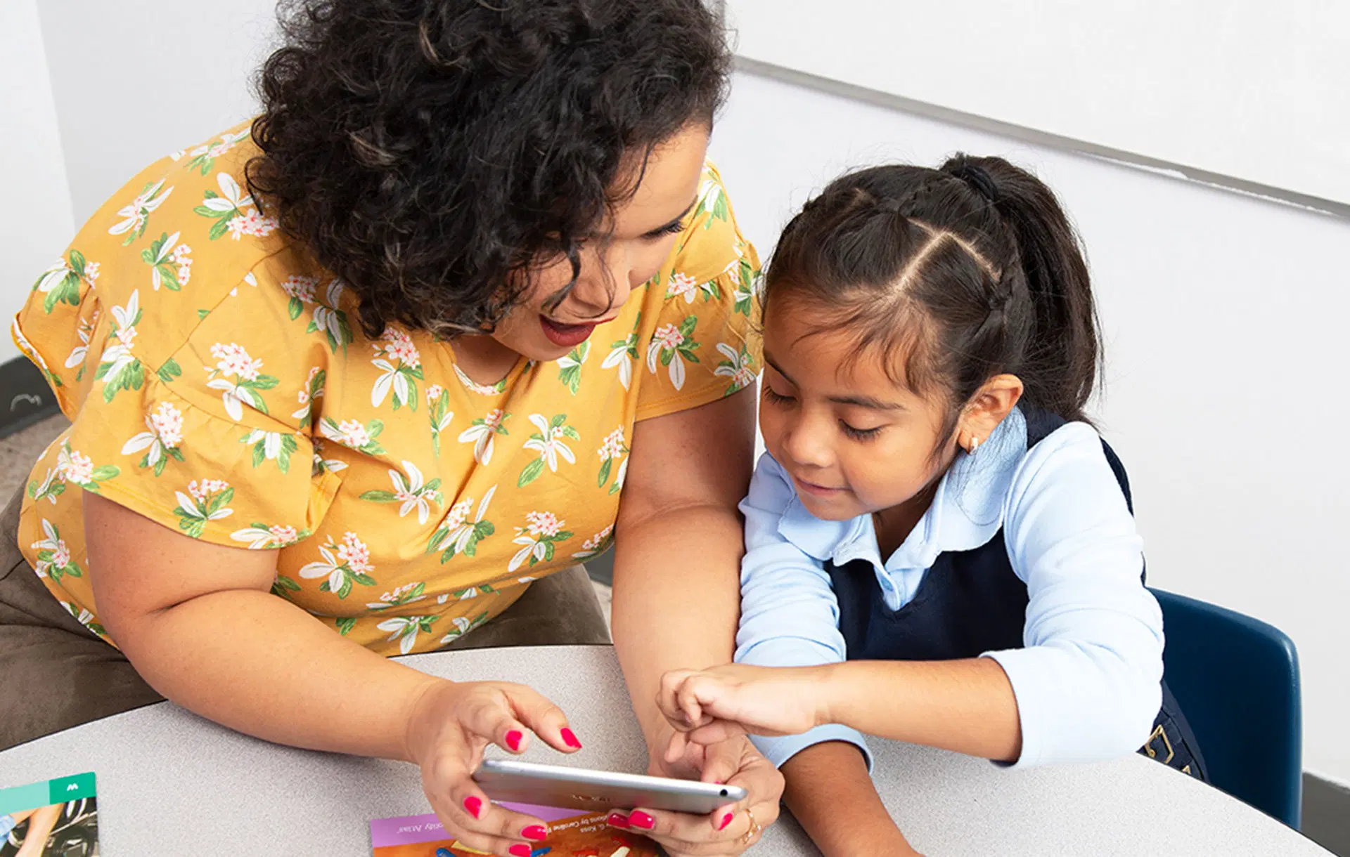 Un adulto y un niño se sientan en una mesa y usan juntos una tableta. El adulto viste un top floral amarillo y el niño, vestido con un traje azul marino y blanco, señala la pantalla.