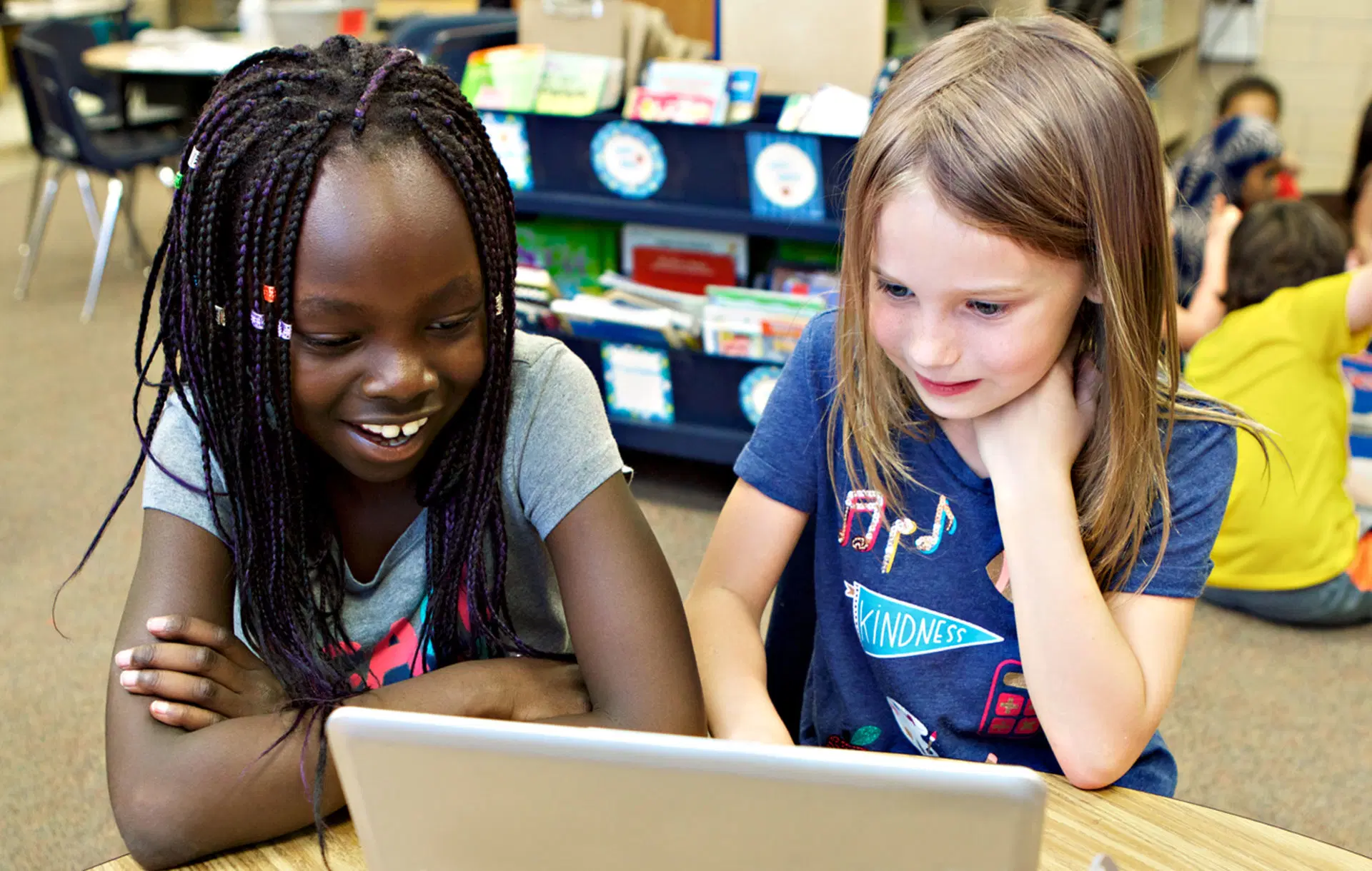 Dos niñas están sentadas en una mesa, mirando juntas la pantalla de una computadora portátil en un salón de clases. Al fondo se ven estanterías con libros y materiales de clase.