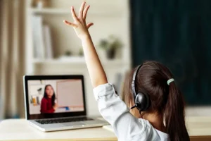 A child wearing headphones raises their hand while attending an online class on a laptop, engaging with resources from the amplify curriculum.