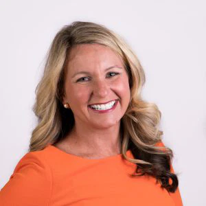 A smiling woman with shoulder-length blonde hair, wearing an orange blouse and gold earrings, against a light background.