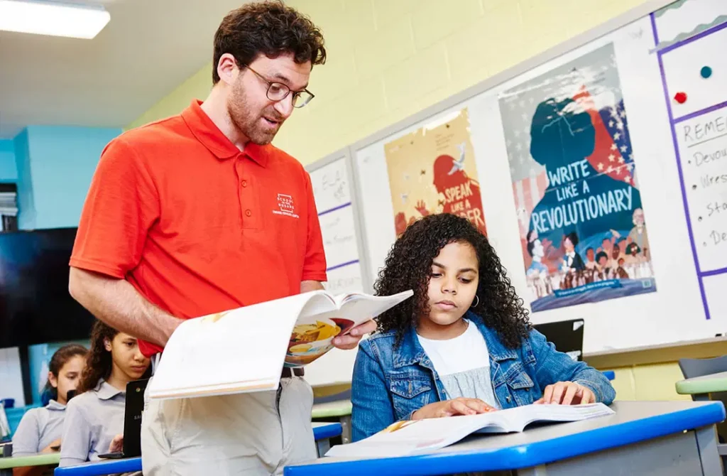 A male teacher in a red shirt explaining a lesson to a young female student who is reading a book in a classroom.