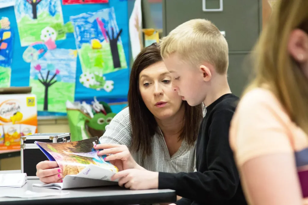 A woman and a young boy looking at a book together in a classroom, with colorful artwork displayed in the background.