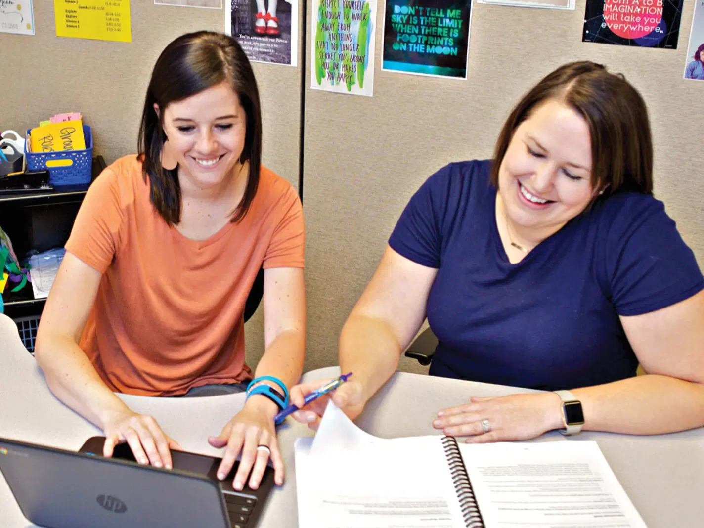 Two women sit at a desk; one types on a laptop while the other writes in a spiral notebook. Both are smiling. Colorful posters are visible on the wall behind them.