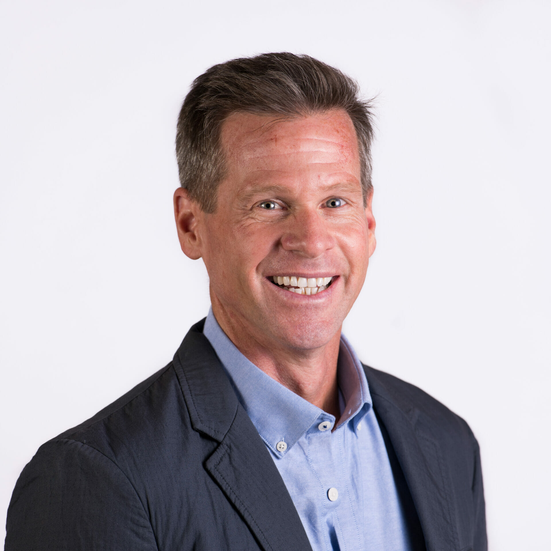 Un hombre con cabello castaño corto sonríe a la cámara. Lleva una camisa azul con botones y una chaqueta oscura. El fondo es completamente blanco.
