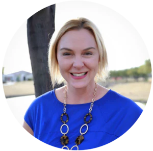 A smiling person with short blonde hair is wearing a blue top and a large, circular-link necklace, standing outdoors with trees and grass in the background.
