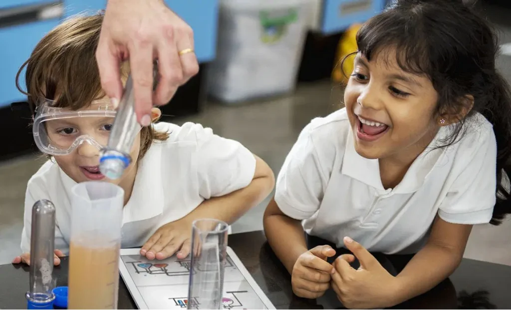 Dos niños con polos blancos observan a una persona verter líquido de un tubo de ensayo en una probeta graduada. Un niño usa gafas de seguridad y parece emocionado, mientras que el otro sonríe y señala.