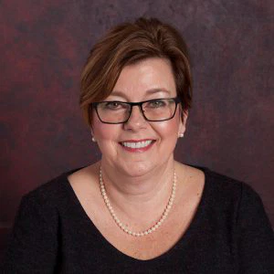 Portrait of a smiling woman with short brown hair, wearing glasses, a black blouse, and a pearl necklace against a maroon background.