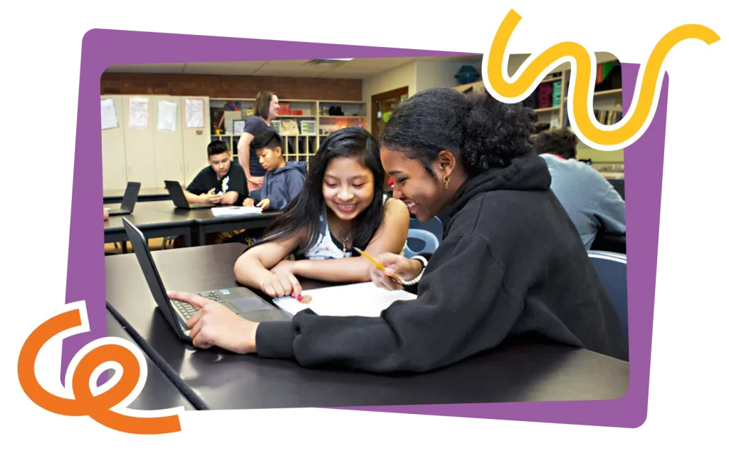 Two students, one black and one asian, smiling and studying together at a laptop in a classroom with other students in background.