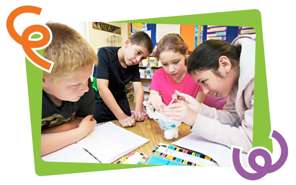 Four children around a table engaged in a classroom science project, examining materials closely, with educational posters in the background.