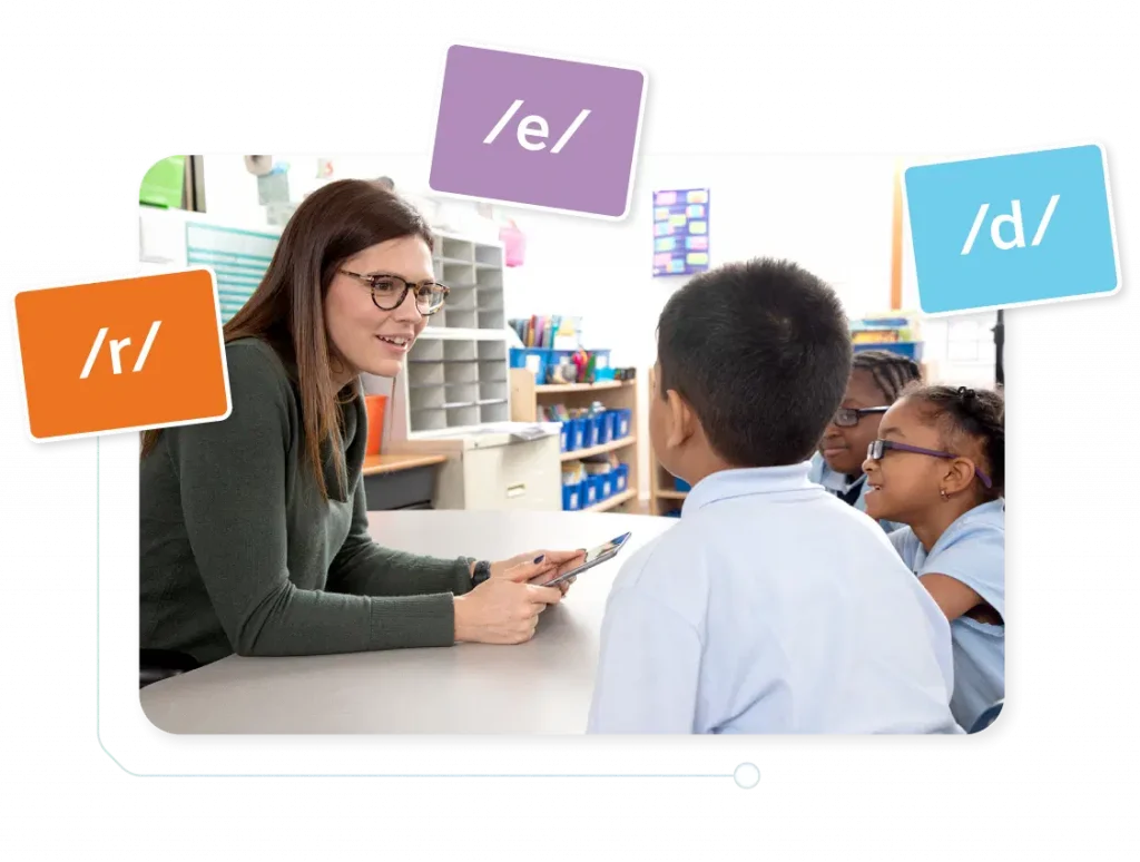 A teacher interacts with three young students at a desk, using flashcards to teach phonetics in a classroom.