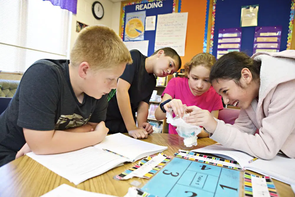 Cuatro niños reunidos alrededor de una mesa en un salón de clases, mirando de cerca un objeto que uno de ellos sostiene. Hay libros, papeles y materiales educativos sobre la mesa.