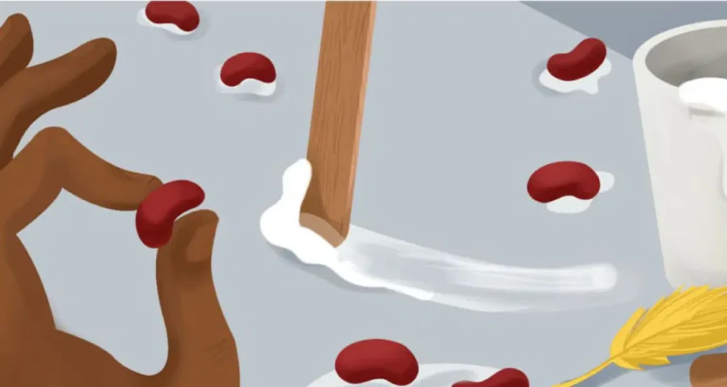 A hand holds a red bean on a table, while a wooden stick spreads white glue and scattered beans. Also on the table are a white cup and a yellow pen.