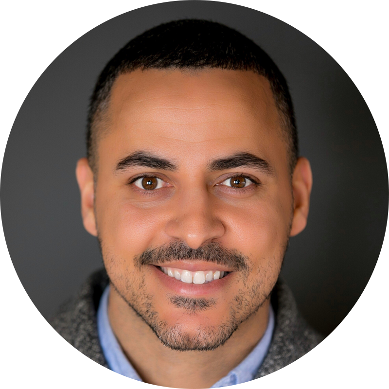 Primer plano retrato de un hombre con pelo corto oscuro y barba recortada, sonriente, vestido con un suéter gris sobre una camisa azul. El fondo es gris oscuro.