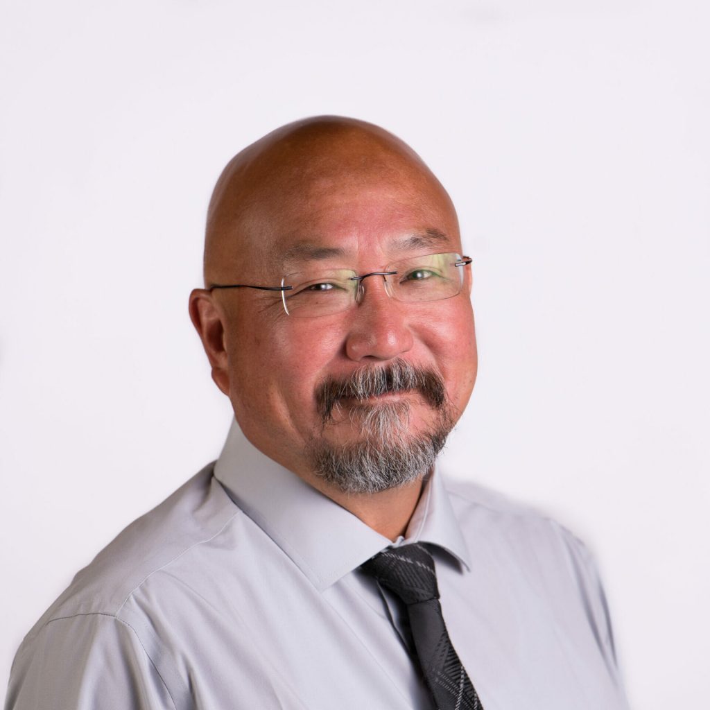 A middle-aged man with glasses, a mustache, and a goatee, wearing a light gray shirt and a black tie, posed against a plain white background.