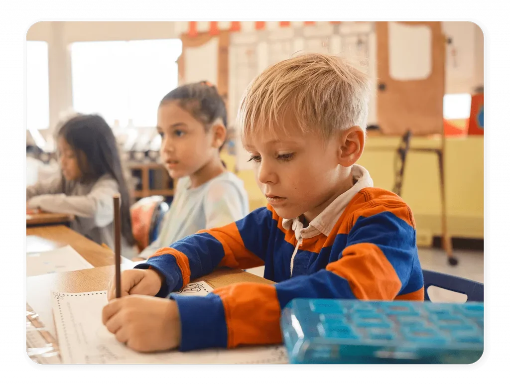Young boy in a classroom sits at a desk and writes on a worksheet, with other children working at nearby desks in the background.