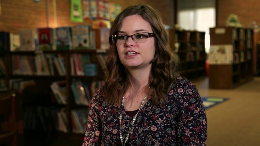 A woman with glasses and wavy hair sits in a library with bookshelves in the background.