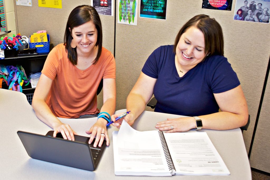 Two women sit at a table working together. One types on a laptop while the other writes on a spiral-bound notebook. Posters and office supplies are in the background.