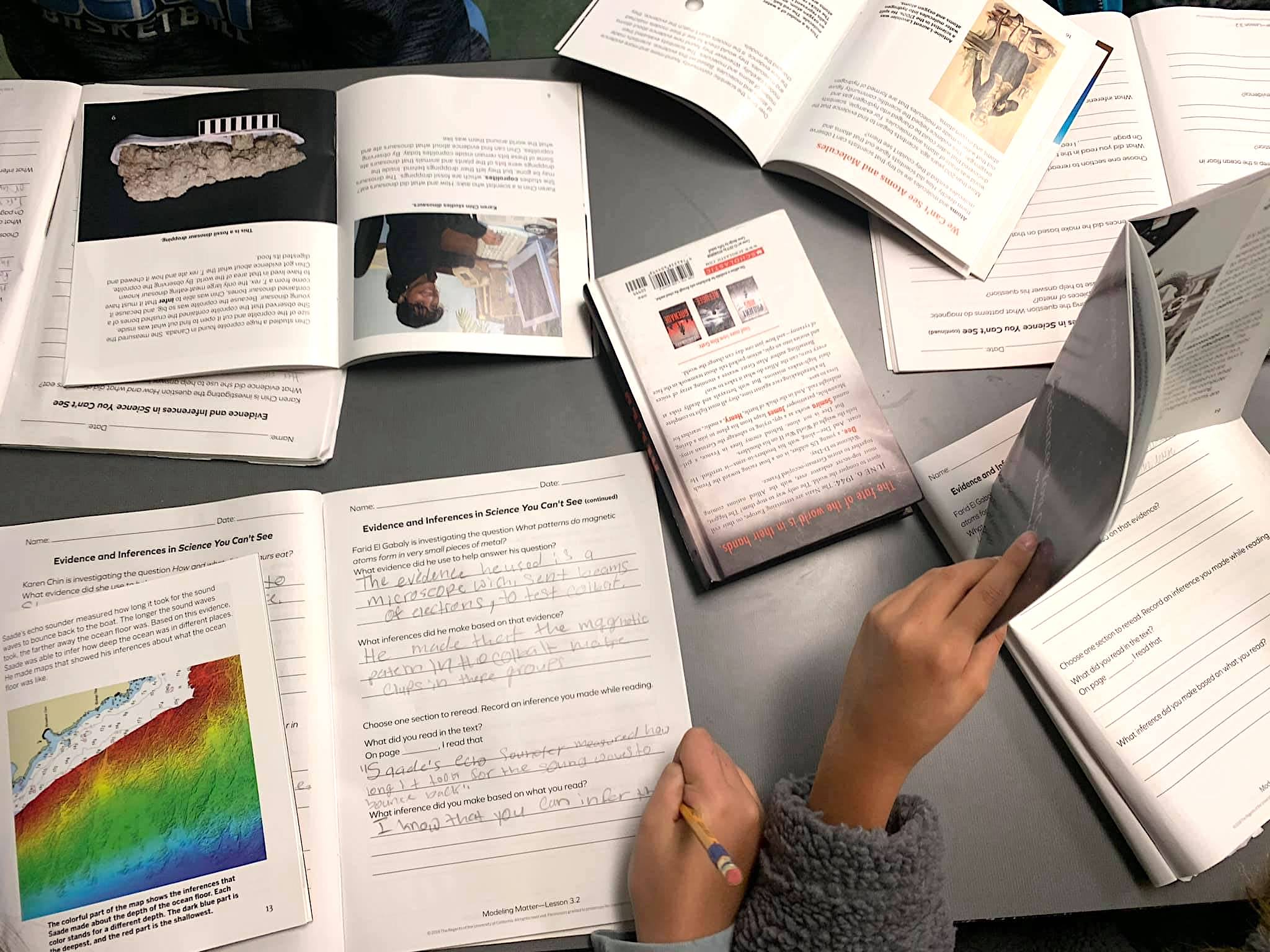 Students working on an assignment at a desk surrounded by open textbooks and notebooks. A person's hands are visible, holding a book and writing with a pen.