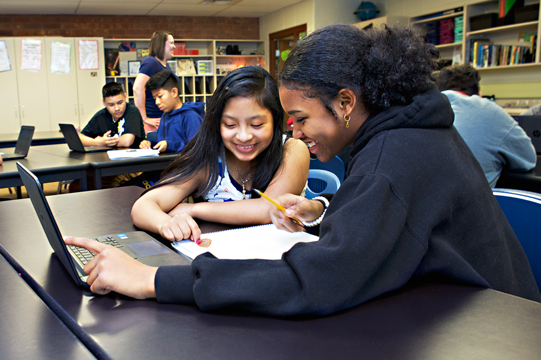 Two students collaborate on schoolwork at a table, with a laptop and a notebook. Other students are focused on their own tasks in the background.