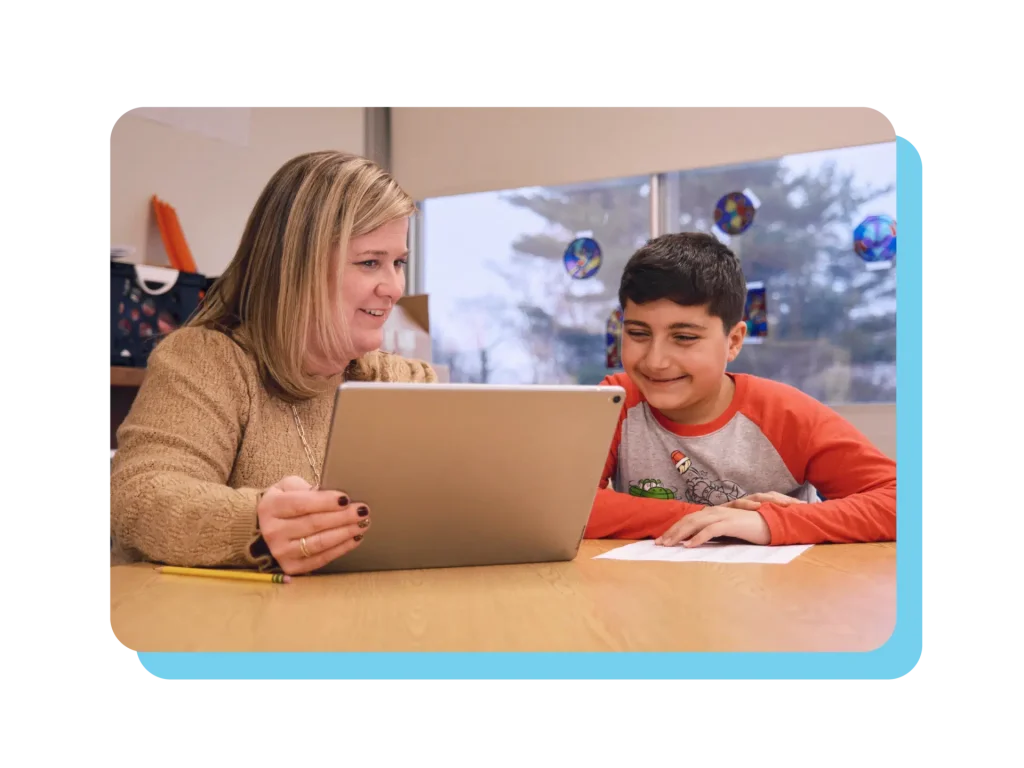 A woman and a boy sit at a table, exploring reading competency on a laptop. The woman holds a pencil, and the boy smiles brightly. Papers are scattered around, with a window adorned with circular decorations in the background.