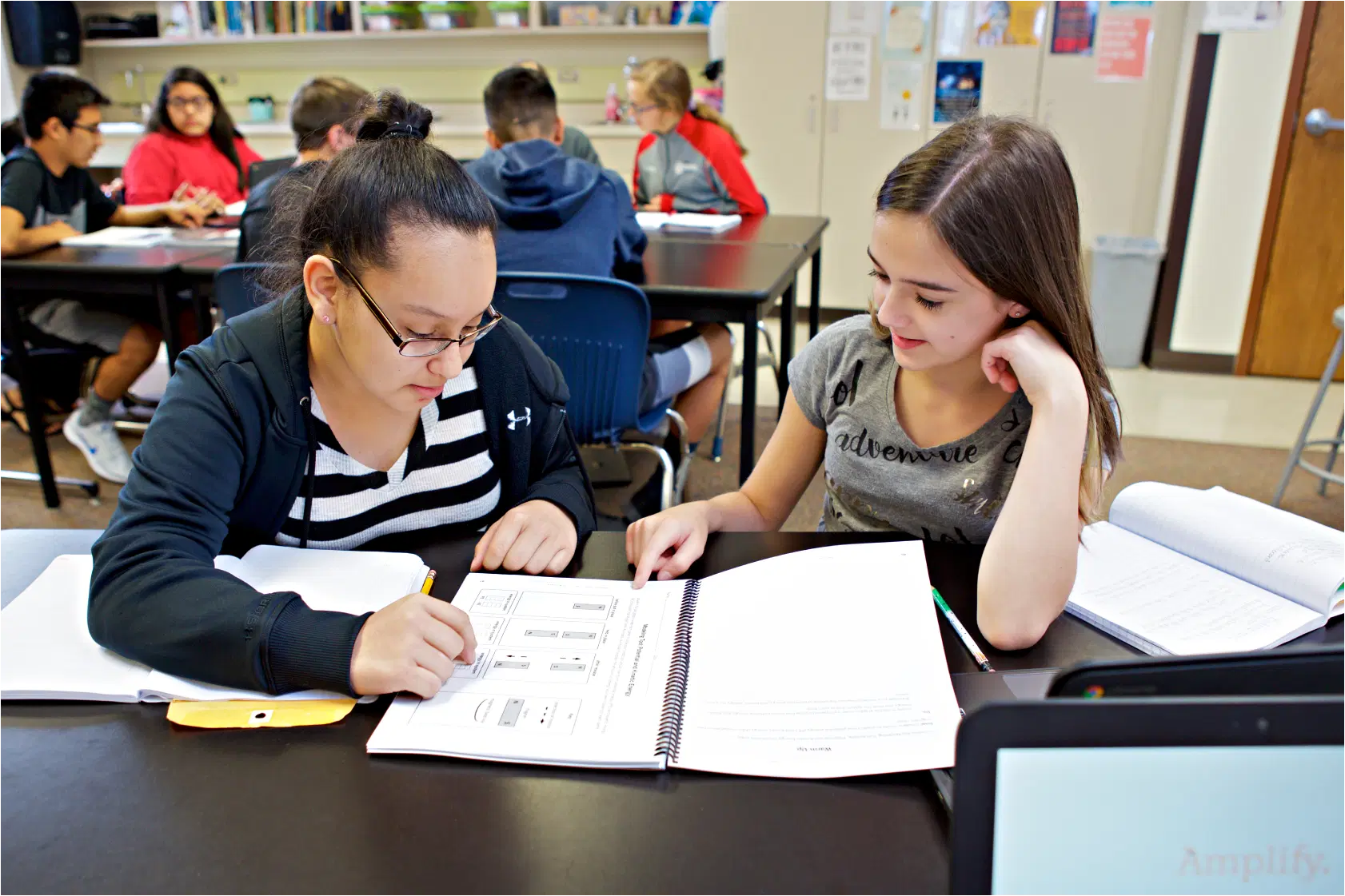 Two students collaborate at a desk, discussing a science workbook. Other students are visible in the background, engaged in the vibrant atmosphere of the classroom.