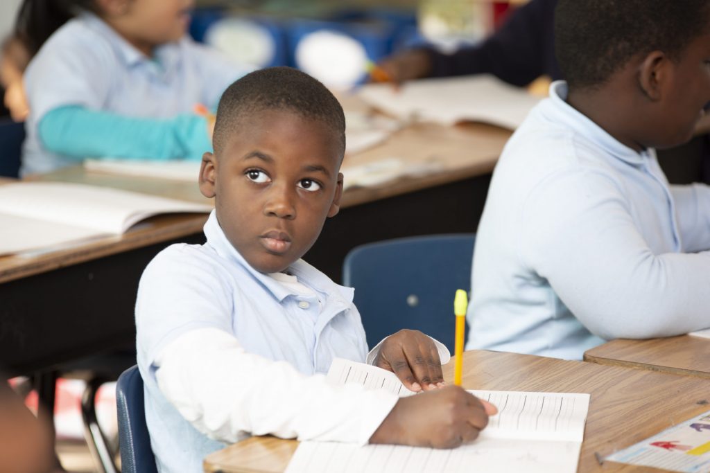 Young student in blue shirt writing in a notebook while sitting at a desk in a classroom, looking forward with focus.
