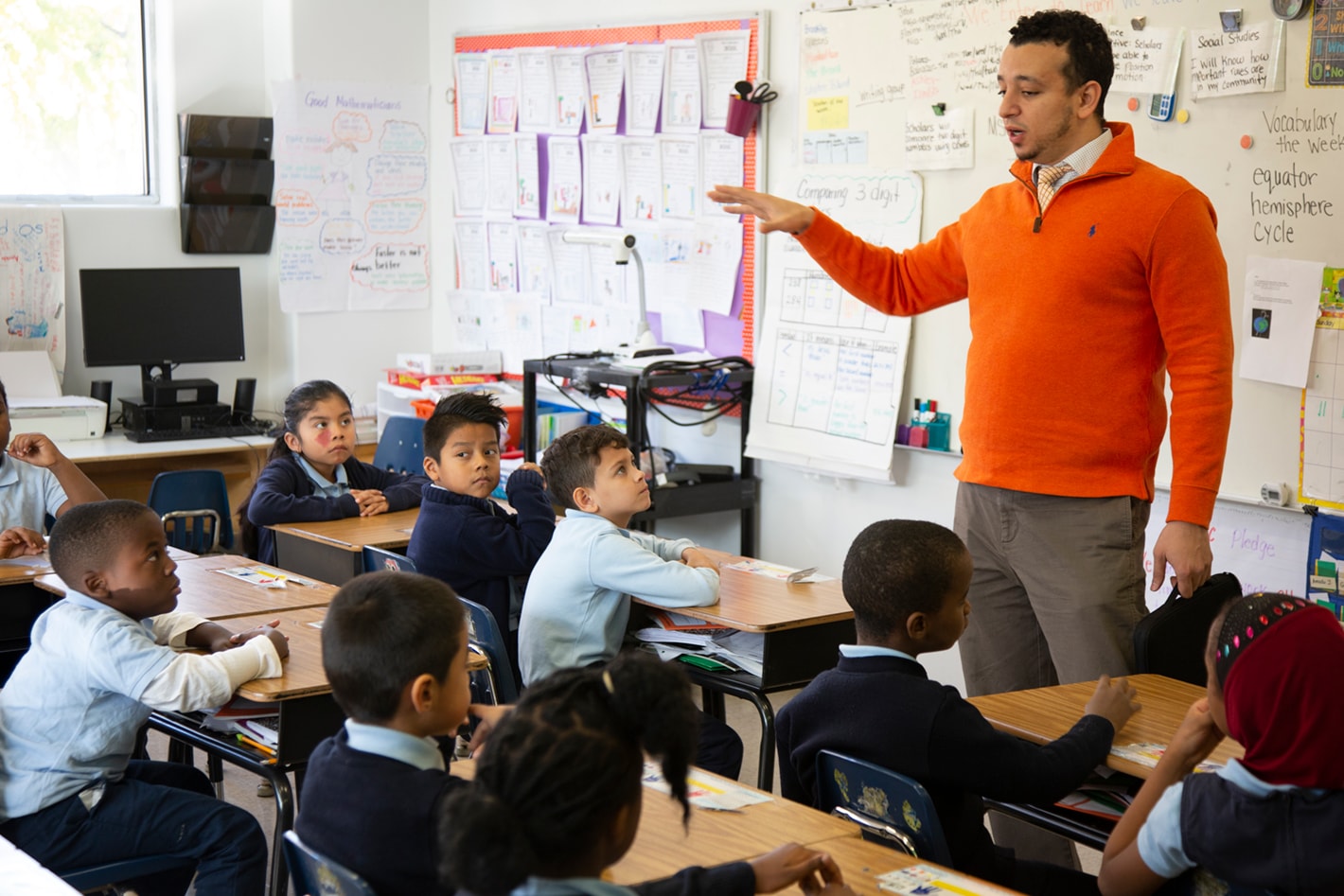 Teacher in an orange sweater gestures while speaking to attentive students seated at desks in a classroom with educational posters on the walls.