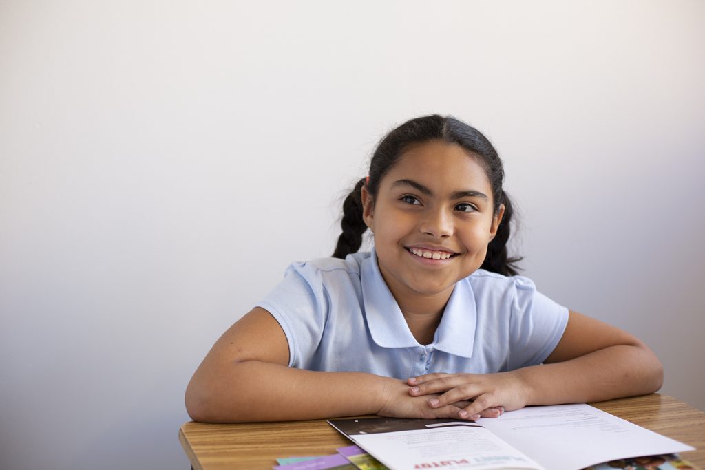 A smiling girl with dark hair in pigtails sits at a desk, looking to the side. She is wearing a light blue shirt and has an open book in front of her.